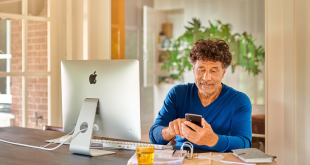 Man van middelbare leeftijd tikt iets in op zijn mobiel terwijl hij achter zijn desktop zit.